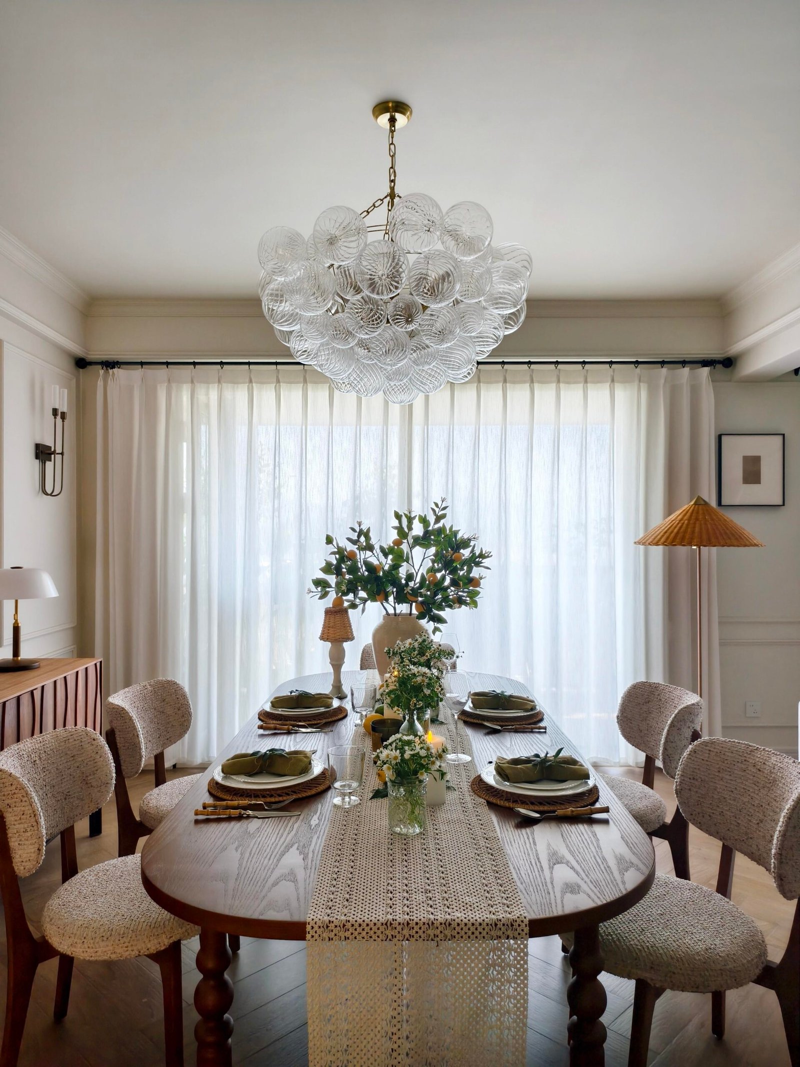 Oval dining room table styled beneath a bubble glass chandelier and sheer curtains